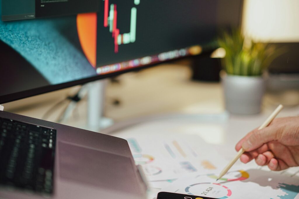 Person notiert Zahlen auf Papier neben einem Monitor bei der Analyse von Finanzdaten